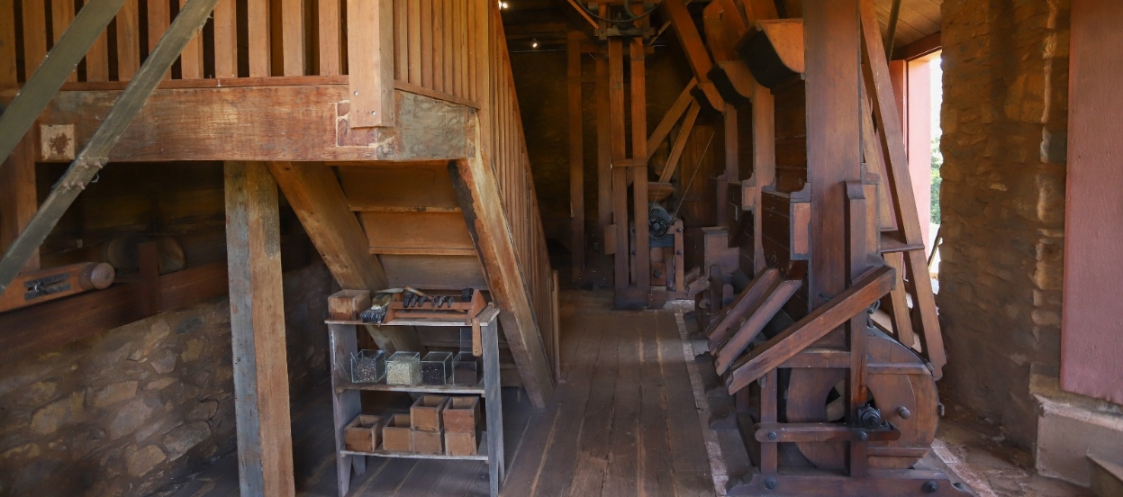 Foto interna da tulha. À esquerda, parte traseira de uma escadaria fixa de madeira, sobre estante com utensílios. À direita e ao fundo, partes da máquina para beneficiar café, do século XIX, majoritariamente em madeira. As paredes laterais e do fundo são de pedras.