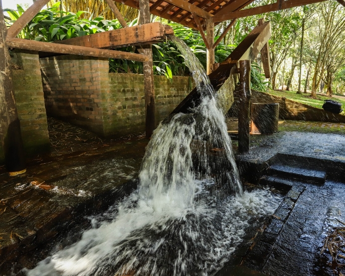 Foto do monjolo (sistema para moer grãos movido pela força da água) em funcionamento. Construído em madeira, está embaixo de uma estrutura com telhado e sem paredes. Ao redor, vegetação.