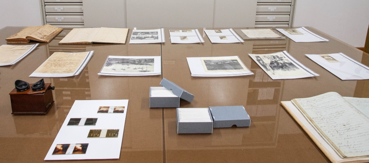 Foto de objetos arrumados sobre uma mesa: fotos e documentos em envelopes de poliéster transparente; três livros abertos; um instrumento óptico e algumas pequenas fotos, provavelmente em suporte de vidro. Sabemos que se trata da mesa do arquivo da Fazenda do Pinhal.