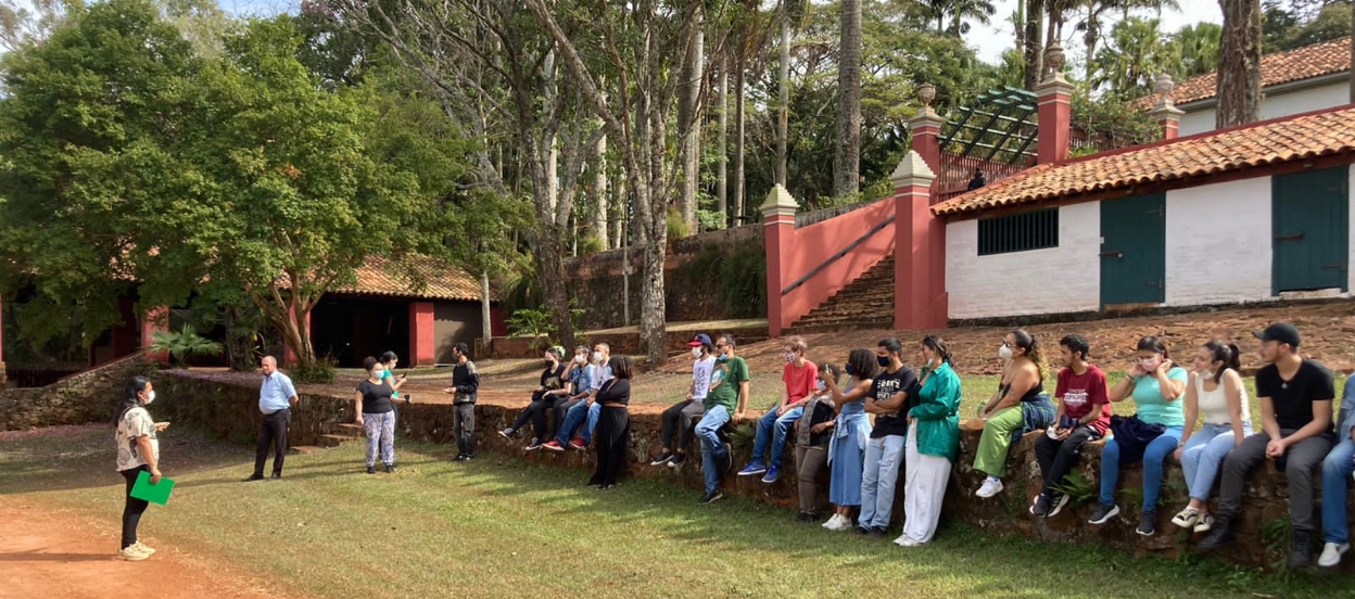 Foto em área externa rural de um grupo de pessoas sentadas em um muro de arrimo de pedras, em frente às cocheiras. Na frente delas, em pé, está uma mulher falando. Ao lado das cocheiras, uma escadaria, no centro da imagem. À esquerda, várias árvores. Sabemos que se trata de uma educadora da Fazenda falando para um grupo de visitantes.