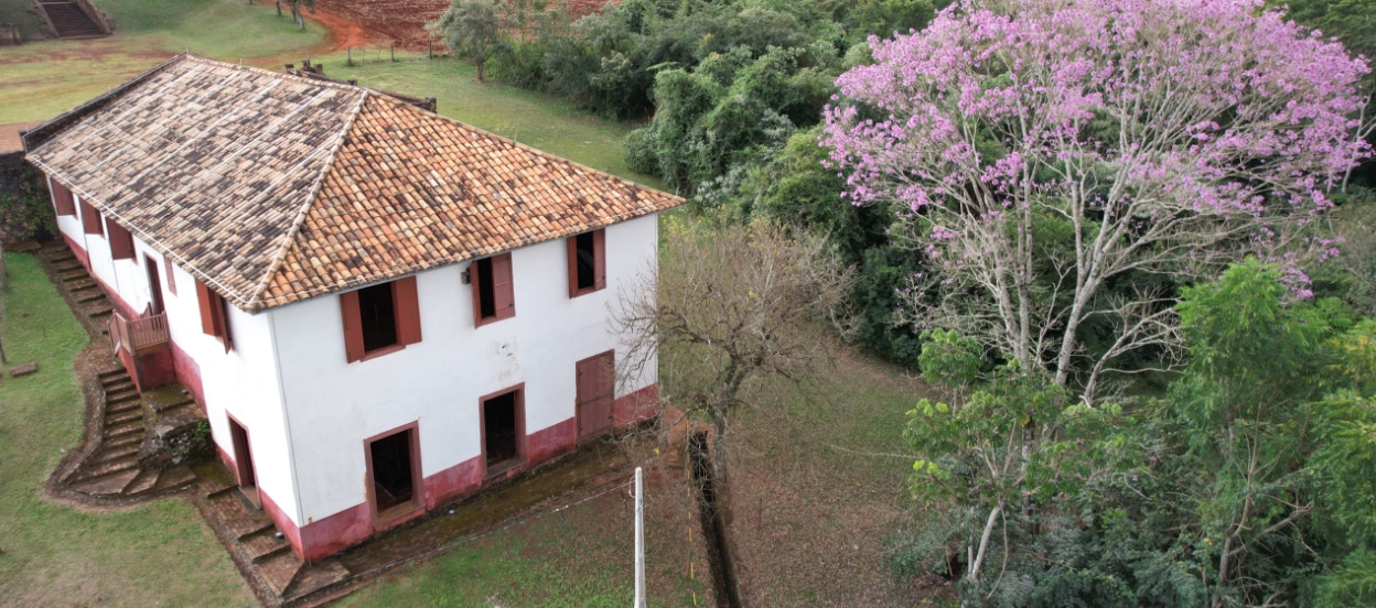 Foto aérea de um grande edifício antigo, a tulha, com três pavimentos (o forro do telhado forma o terceiro). Na fachada frontal, há três janelas no pavimento intermediário e três portas no inferior. Na lateral, uma escada externa acompanha o declive do terreno e há janelas e portas nos diferentes pavimentos. À direita, há árvores, sendo uma com flores cor-de-rosa.