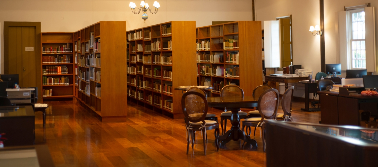 Foto de uma biblioteca com chão de tábuas de madeira. Ao fundo, aparecem três grandes estantes de madeira. Todas contêm livros nas prateleiras. Nas laterais, há mesas encostadas nas paredes. Em frente às estantes, uma mesa oval com cadeiras, para reunião. Sabemos que é a biblioteca do CEPE - Centro de Estudos, Pesquisa e Educação.