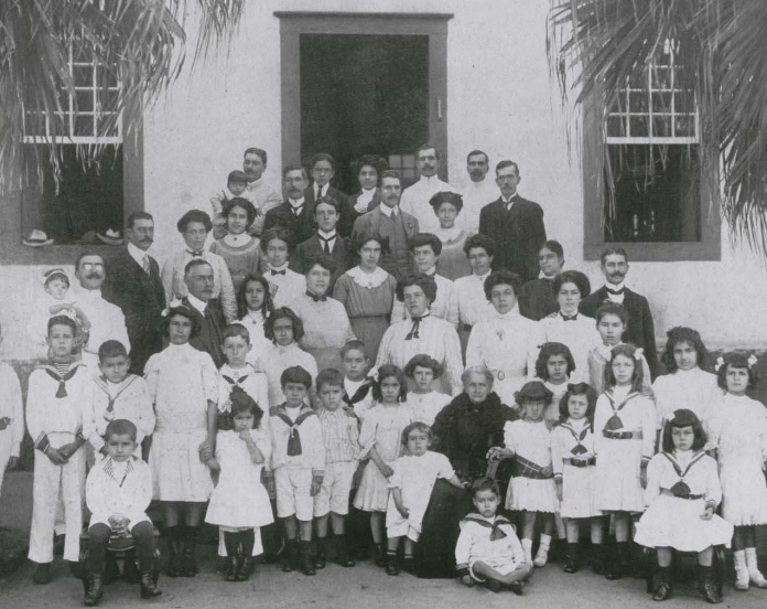 Foto antiga de família, em preto e branco, em escadaria externa, na frente de um casarão. Em pé, várias mulheres e homens e, na frente, muitas crianças, a maioria também em pé. Sentada, de preto, há uma senhora já idosa. Sabemos que se trata da família Arruda Botelho, no jardim das camaradinhas, e que a senhora de preto é a condessa do Pinhal. A foto é de junho de 1910, de autoria de Guilherme Gaensly.