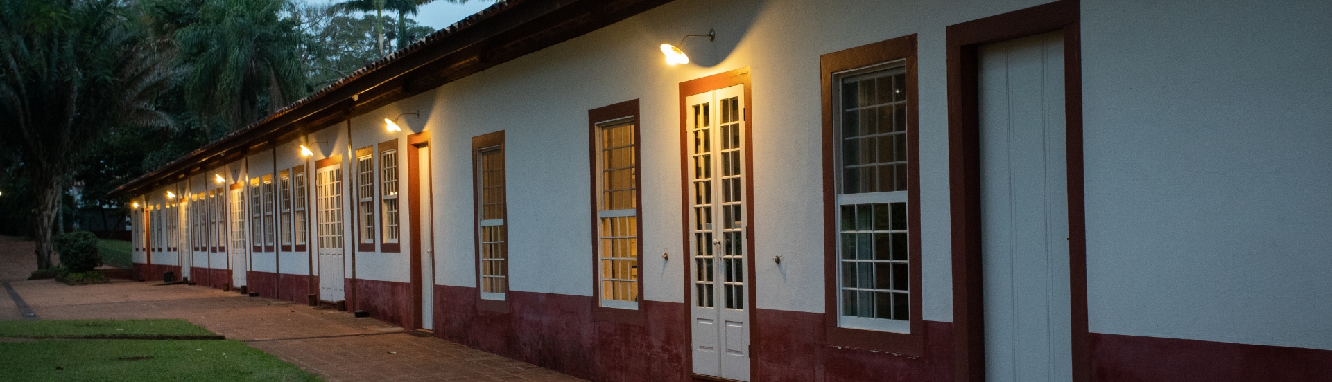 Foto externa, no anoitecer, de uma edificação alongada com diversas portas e janelas. A parede é branca com um barrado em vermelho queimado, mesma cor que emoldura as portas e janelas. Há luzes acesas acima das portas. Ao fundo, há árvores. Sabemos que se trata da antiga senzala.