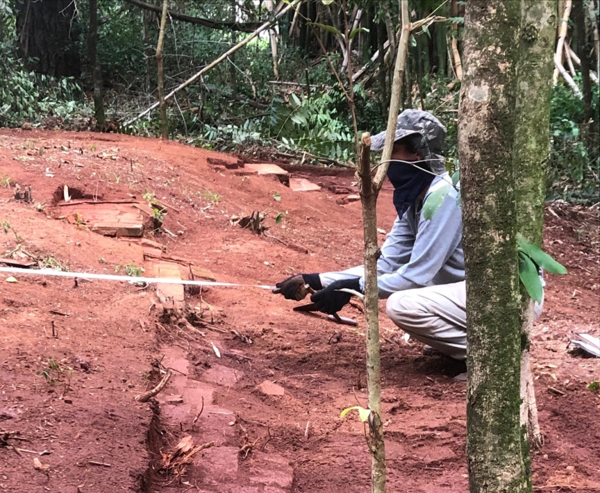 Foto de uma pessoa de perfil, agachada, vestindo blusa, calças compridas, luvas, chapéu e lenço cobrindo o rosto, aparentemente na clareira de uma área de mata. Ela segura uma fita de medição esticada na horizontal, em um terreno de terra vermelha levemente inclinado. Sabemos que se trata de um arqueólogo em atividade na Fazenda do Pinhal, no ano de 2023.