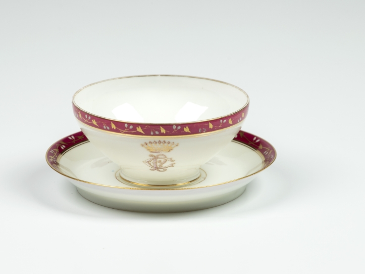 Foto de xícara antiga, sobre pires em fundo brando. A louça é branca, com uma faixa vermelha nas bordas, ornada com ramos dourados e um friso dourado. Na xícara, um monograma dourado com as letras C e P sobrepostas sob uma coroa. Sabemos que faz parte do jogo de jantar de Antonio Carlos de Arruda Botelho, conde do Pinhal.