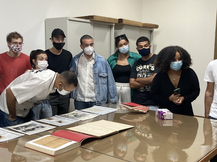 Foto de visitantes, em pé, ao redor de uma mesa onde há alguns livros abertos, outros empilhados; fotos e documentos em envelopes de poliéster transparente. Eles olham para alguém (que não aparece na foto). Um homem está abaixado, olhando detidamente para uma foto. Todas as pessoas usam máscaras cobrindo bocas e narizes. Sabemos que se trata de visita ao arquivo da Fazenda do Pinhal, quando ainda havia perigo de contaminação de coronavírus, em 2022.